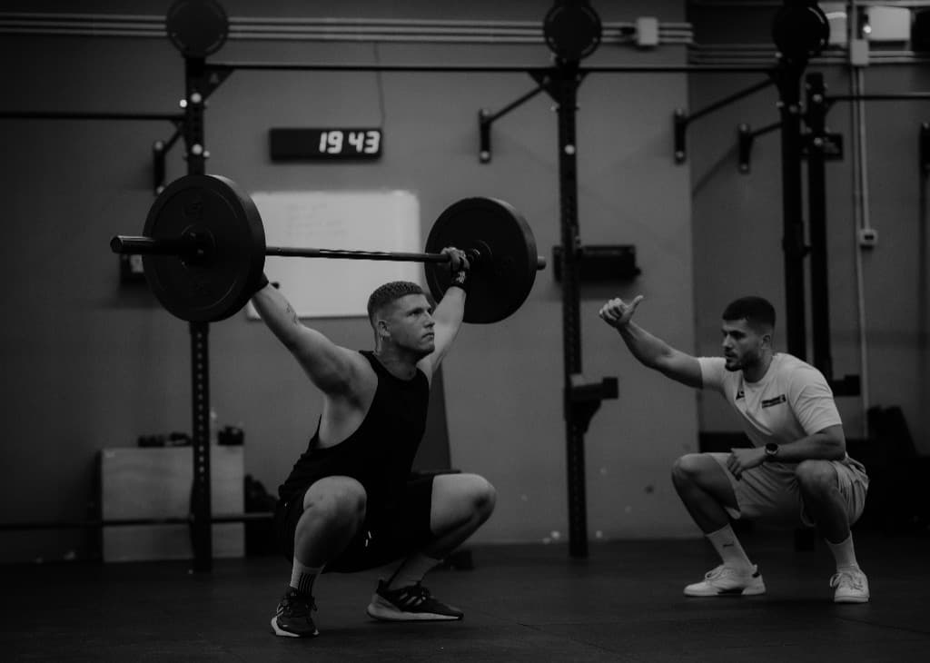 Coach giving a thumbs-up while a client holds an overhead squat in a gym